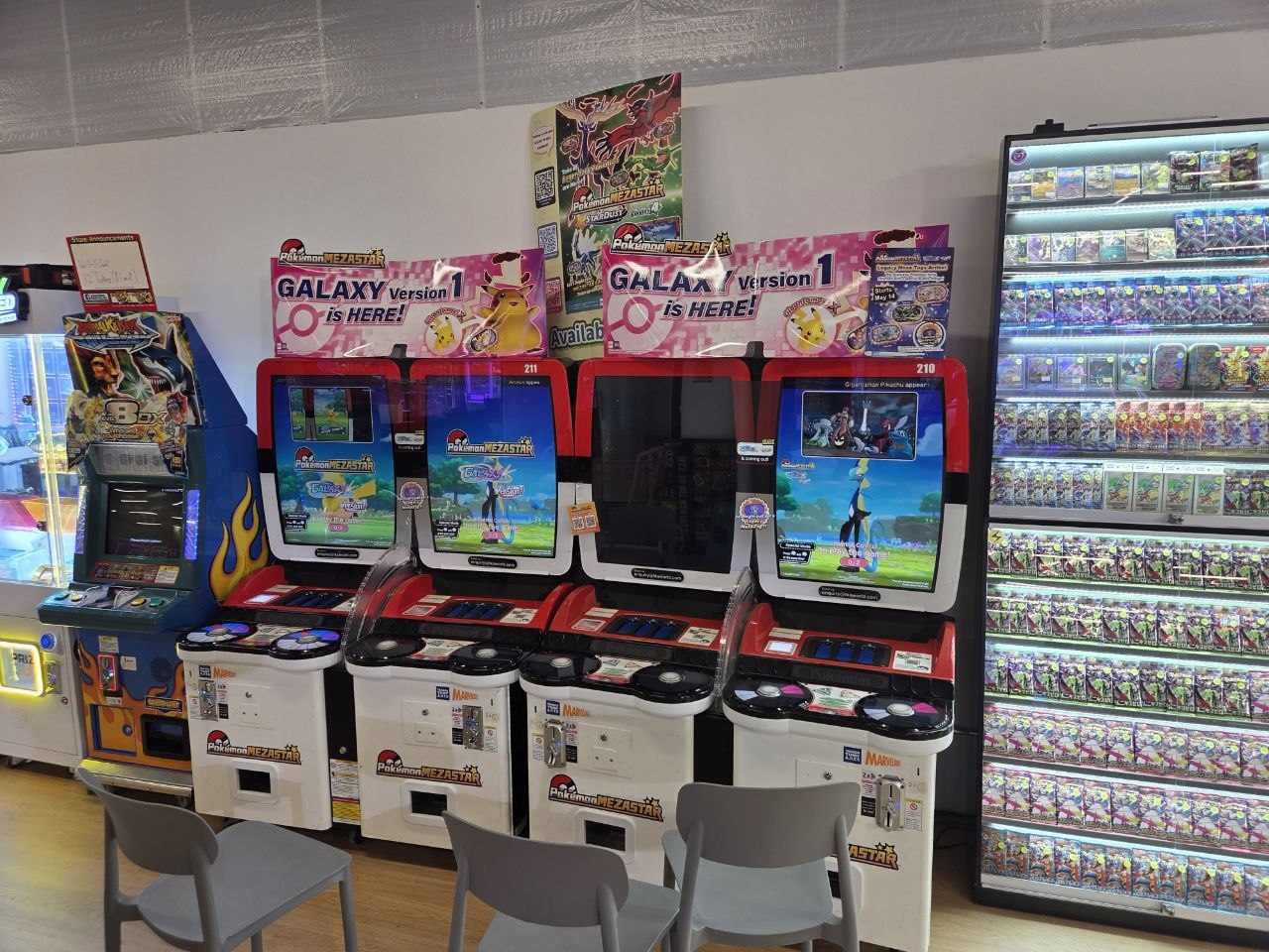 Pokemon Mezastar arcade cabinets at Play United with grey chairs in front for seating, beside a tall lightbox display wall of Japanese Pokemon booster packs