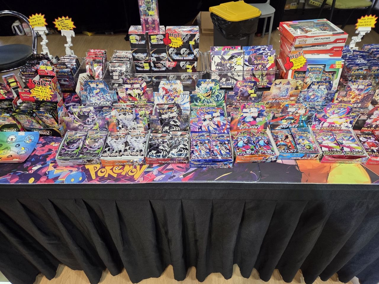 A table at Play United covered with sealed Japanese Pokemon booster boxes and packs marked with Sale and Hot price tags, with a Pokemon-branded table cloth